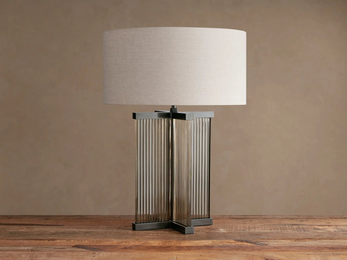 Delsie Table Lamp with a flax fabric drum shade over a fluted glass and dark bronze steel base on a rustic wooden table.