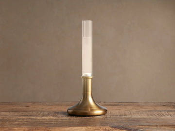 Luella Outdoor Rechargeable Lamp: burnished brass base & clear glass tube with glowing LED, standing on a rustic wood table.