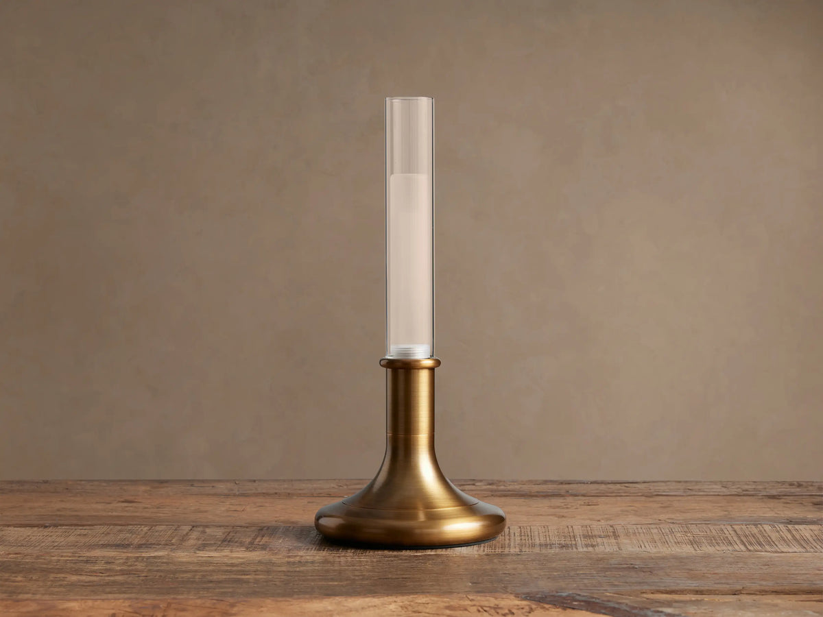Luella Outdoor Rechargeable Lamp: burnished brass base & clear glass tube with glowing LED, standing on a rustic wood table.