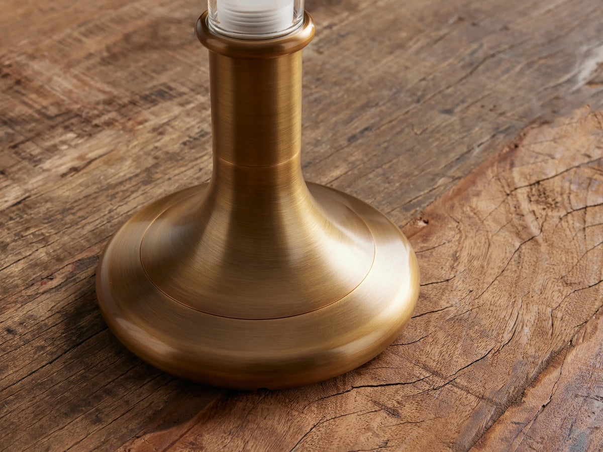 Luella Outdoor Rechargeable Lamp with a burnished brass base & clear glass cylinder on a wooden table, against a brown wall.