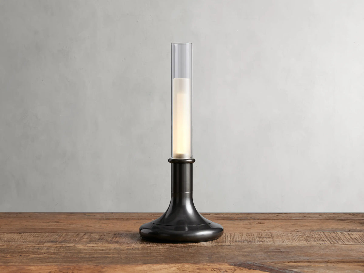 Luella Outdoor Rechargeable Lamp with brushed bronze finish, cylindrical glass top, glowing LED, on wooden table, grey background.
