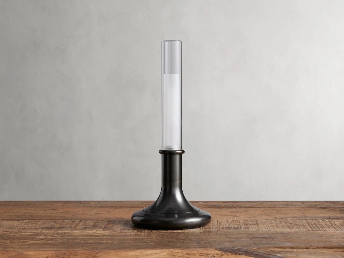 Luella Outdoor Rechargeable Lamp with brushed bronze finish, cylindrical glass top, glowing LED, on wooden table, grey background.