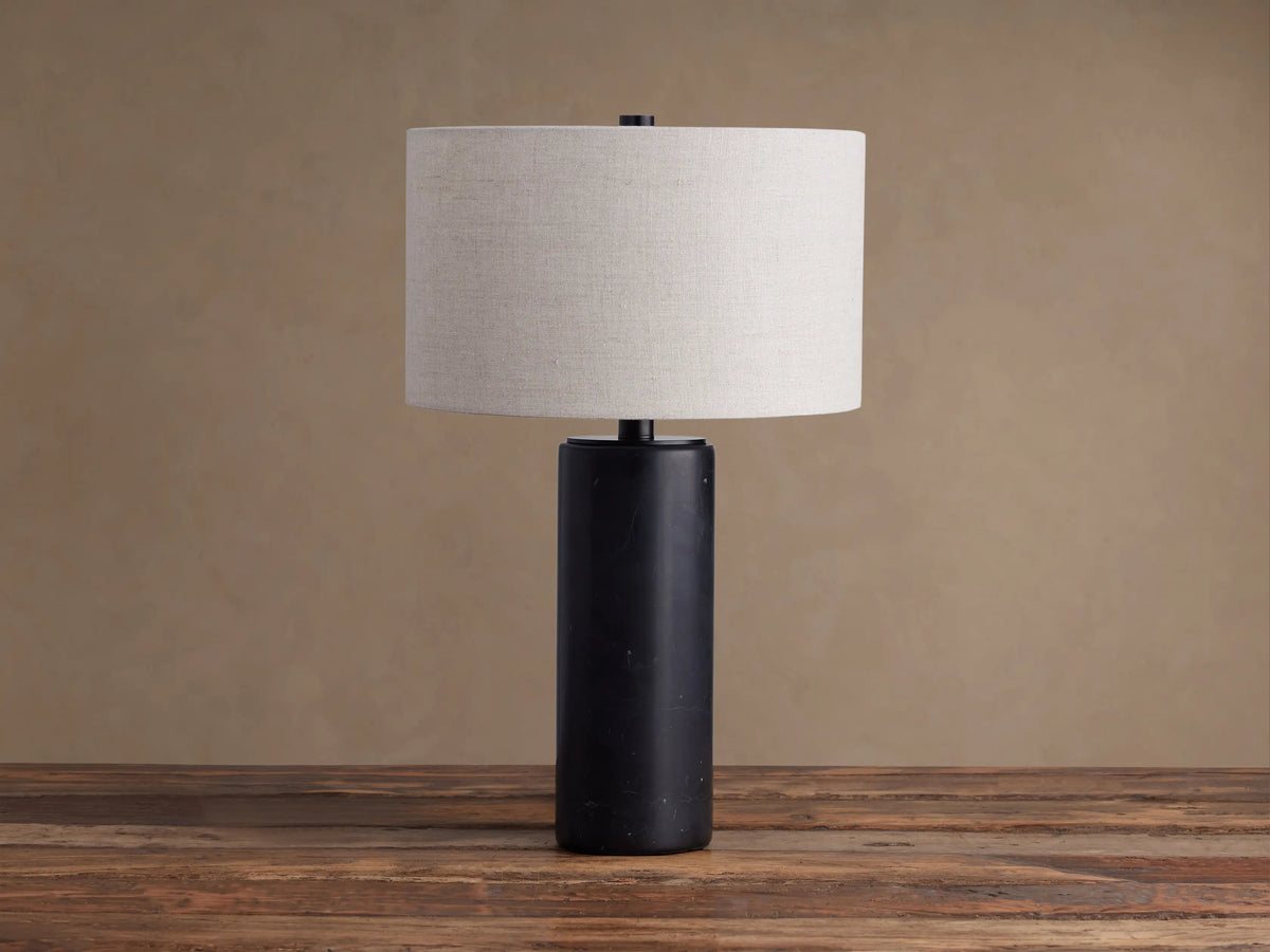 Brixton Table Lamp with a dark, cylindrical marble base & light fabric drum shade on a rustic wooden table.