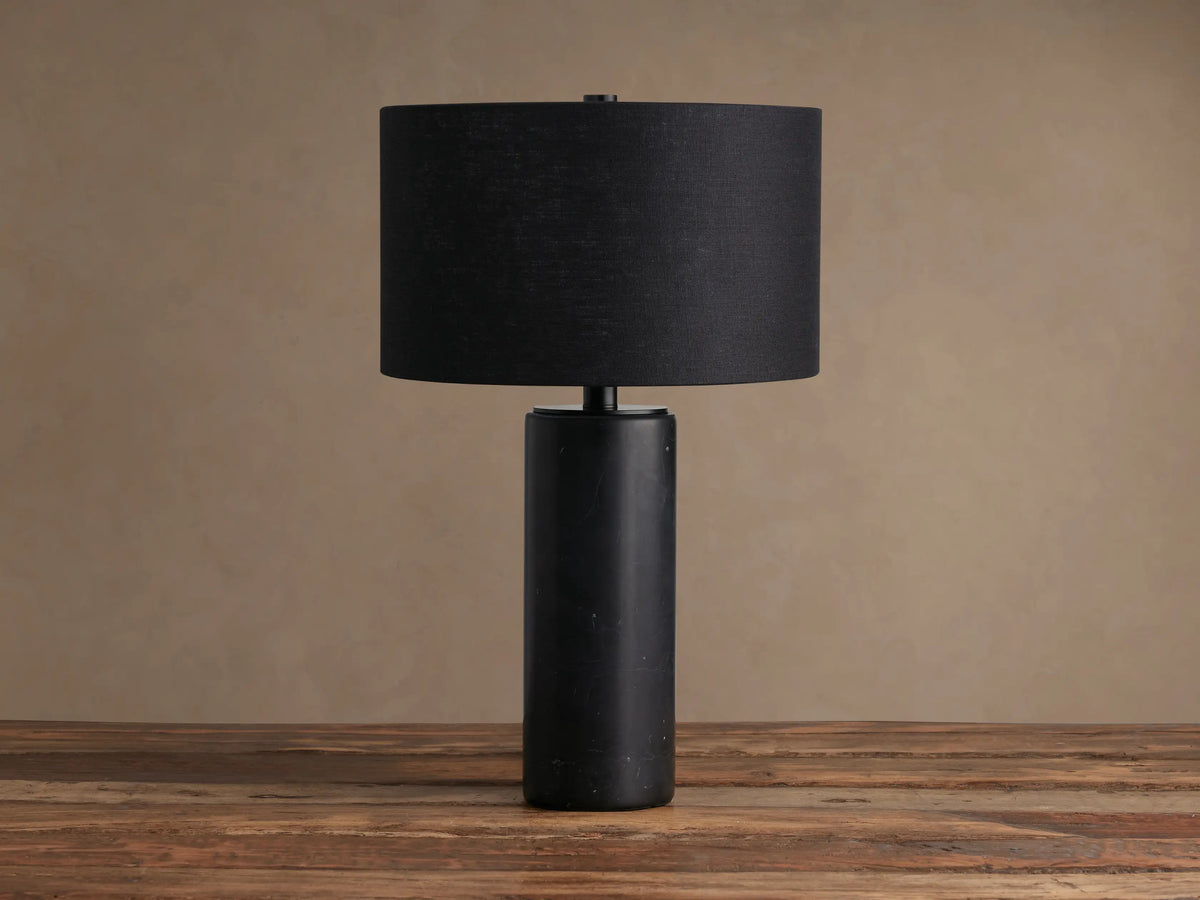 Brixton Table Lamp with a dark cylindrical marble base and a black fabric drum shade, standing on a rustic wooden table.