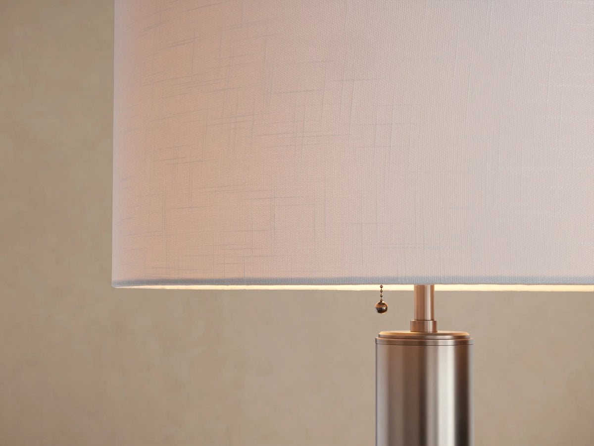 Linden Floor Lamp with a slender pewter-finished pole, round base, and a white cylindrical linen shade. Stands on a wood floor.