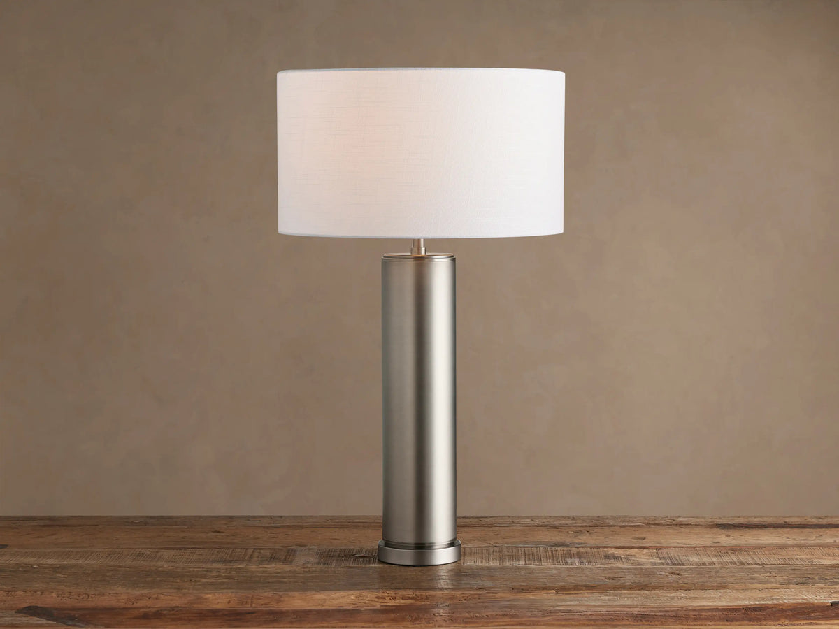 Linden Table Lamp in pewter with cylindrical base, white drum shade, on rustic wooden table against brown wall.
