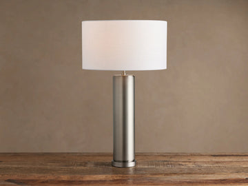 Linden Table Lamp in pewter with cylindrical base, white drum shade, on rustic wooden table against brown wall.