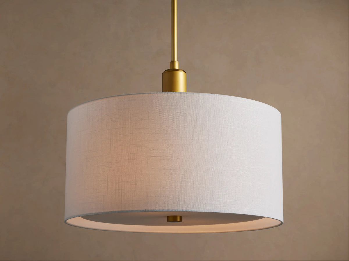 Linden Pendant with a glowing white linen drum shade hanging from a brass rod, seen at a slight angle on a beige background.