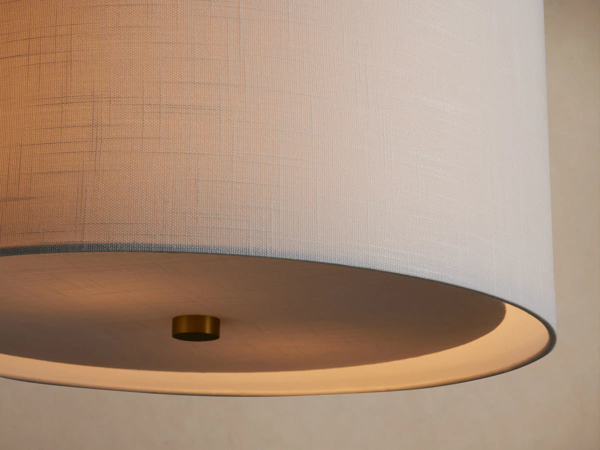 Linden Pendant with a cylindrical light grey linen shade hanging from a brass-finished pole, against a beige background.