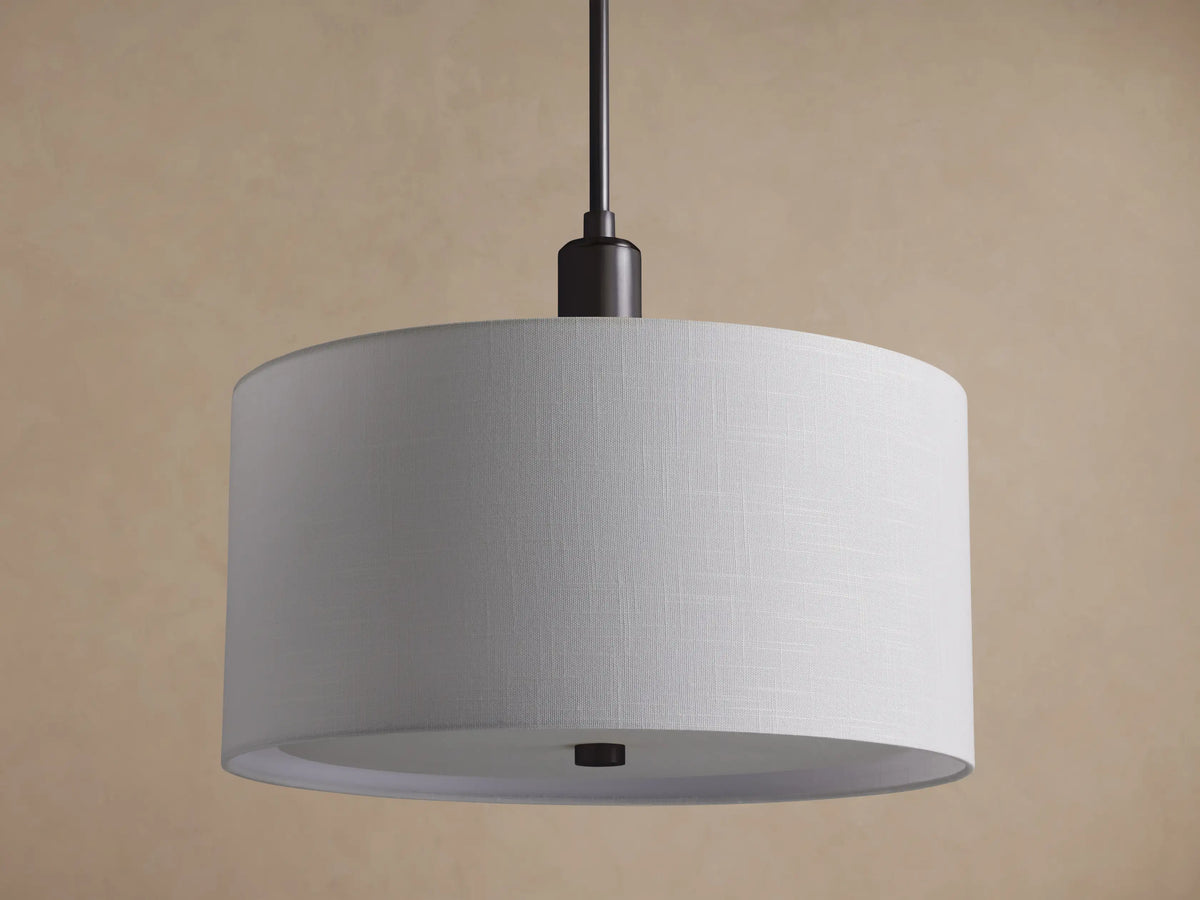 A Linden Pendant hangs, showing its large, round white linen drum shade and dark bronze stem, against a textured brown wall.