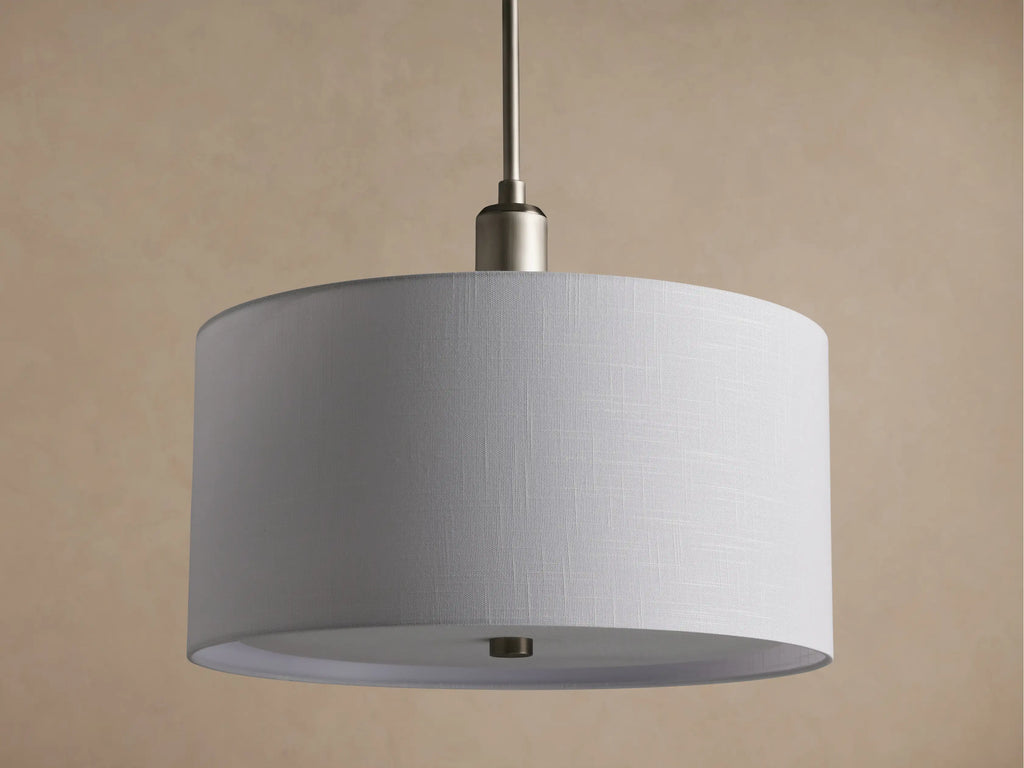 Linden Pendant with cylindrical, off-white linen shade, brushed metallic hardware, glowing softly against a warm wall.