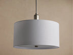 Linden Pendant with cylindrical, off-white linen shade, brushed metallic hardware, glowing softly against a warm wall.