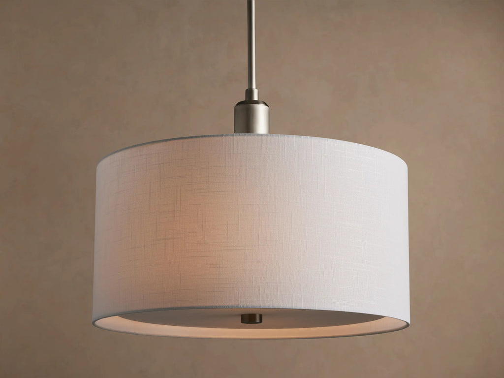 Linden Pendant with cylindrical, off-white linen shade, brushed metallic hardware, glowing softly against a warm wall.