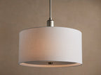 Linden Pendant with cylindrical, off-white linen shade, brushed metallic hardware, glowing softly against a warm wall.