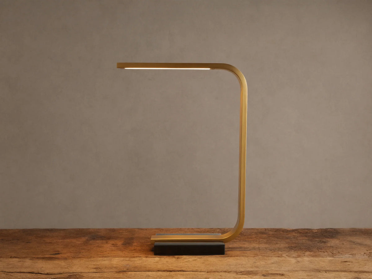 Karson Task Table Lamp: sleek brass, U-shaped lamp with an illuminated LED strip, on a black marble base, resting on a wooden table.