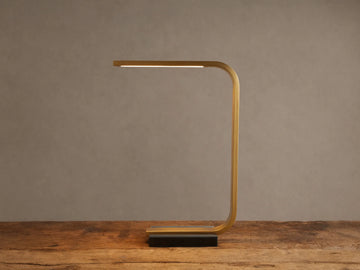 Karson Task Table Lamp: sleek brass, U-shaped lamp with an illuminated LED strip, on a black marble base, resting on a wooden table.