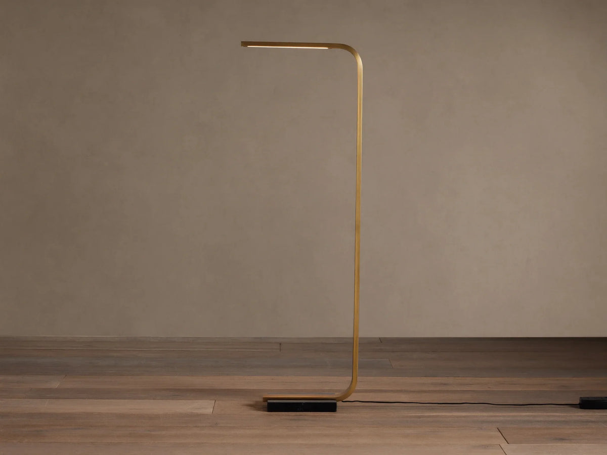 Modern brass Karson Task Floor Lamp with a curved stem, glowing light strip, and black marble base on a wood floor.