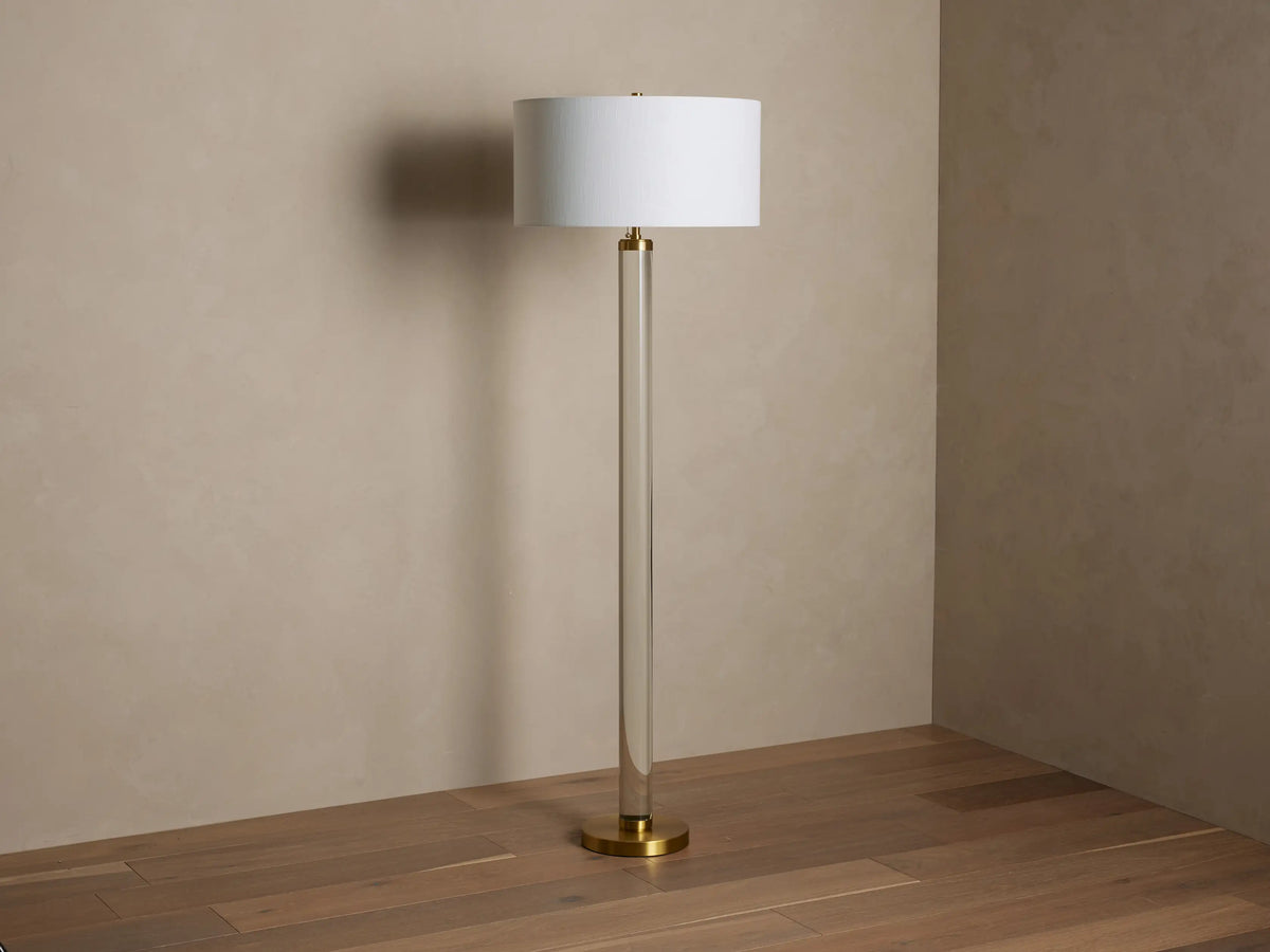 Linden Crystal Floor Lamp with clear column, brass accents, and an illuminated off-white linen shade, on a wood floor.