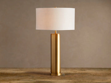 Linden Table Lamp: cylindrical brass-finished iron base, white linen drum shade, on a rustic wood surface.