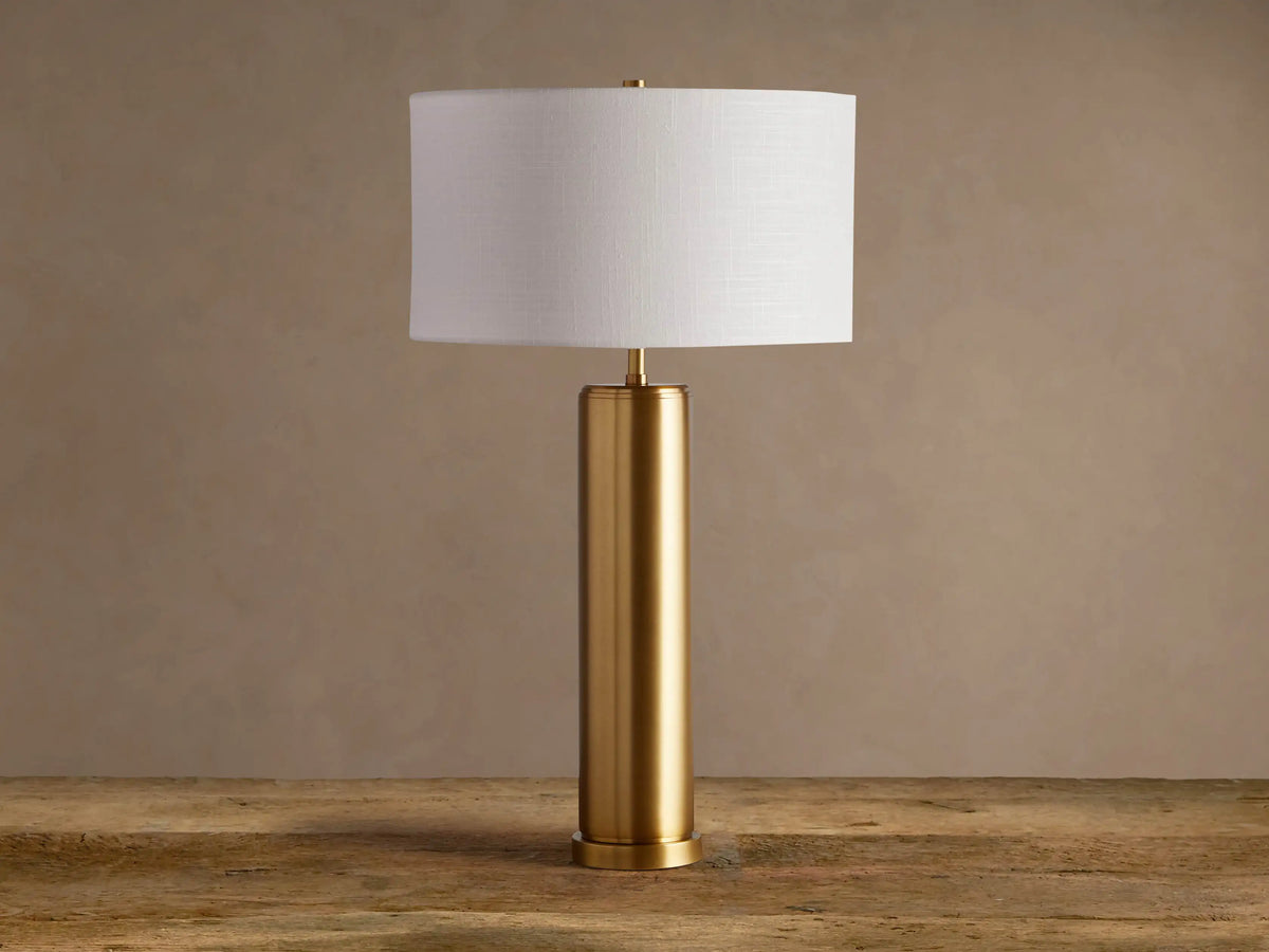 Linden Table Lamp: cylindrical brass-finished iron base, white linen drum shade, on a rustic wood surface.