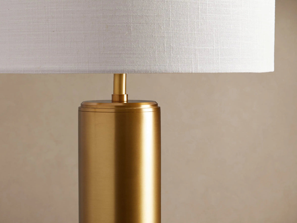 Linden Table Lamp with a polished brass cylindrical base and a white linen drum shade, on a rustic wood table.