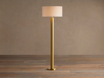 Linden Floor Lamp with a brass-finished pole and round base, topped by a light beige linen drum shade. Standing on a wood floor.