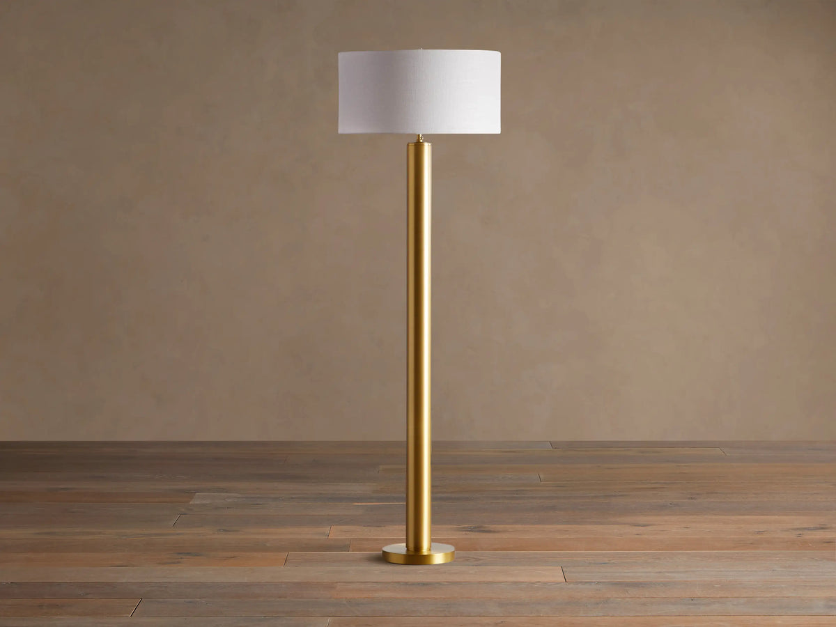 Linden Floor Lamp with a brass-finished pole and round base, topped by a light beige linen drum shade. Standing on a wood floor.