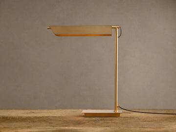 Modern Axle Brass Table Lamp with rectangular shade emitting warm light, slender stand, on rustic wood. Neutral wall background.