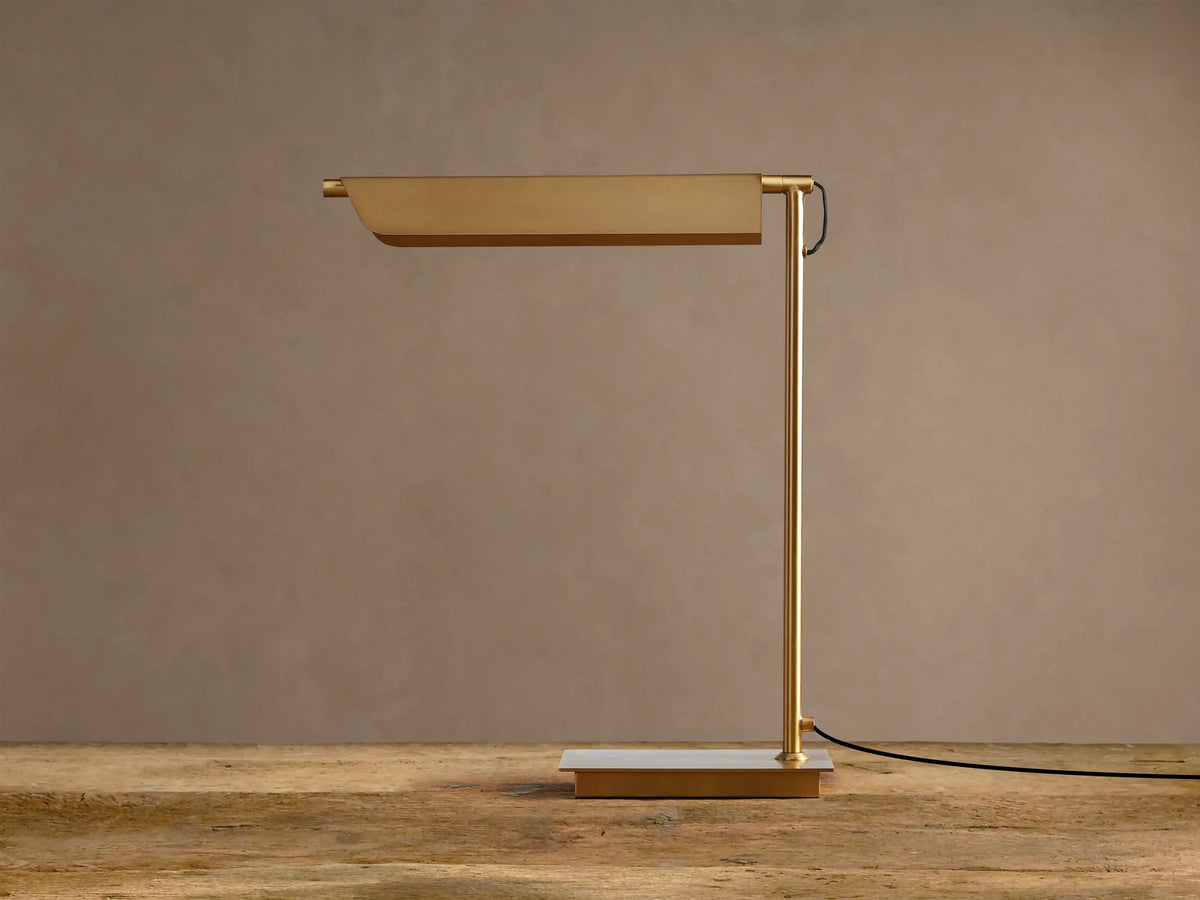 Modern Axle Brass Table Lamp with rectangular shade emitting warm light, slender stand, on rustic wood. Neutral wall background.