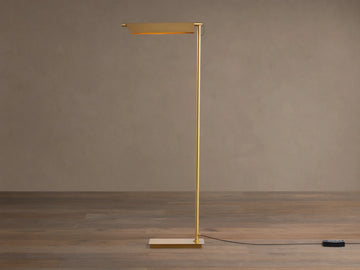 Axle Brass Floor Lamp with a glowing rectangular shade, slim brass pole, square base, and black dimmer on a wooden floor.