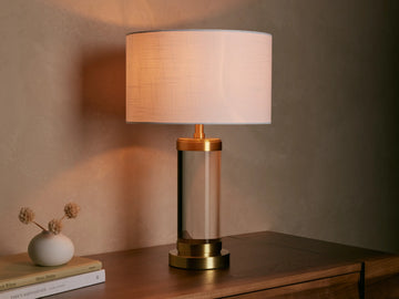 Linden Crystal Table Lamp with clear crystal column, brass accents, and off-white linen shade, glowing on a wooden table.