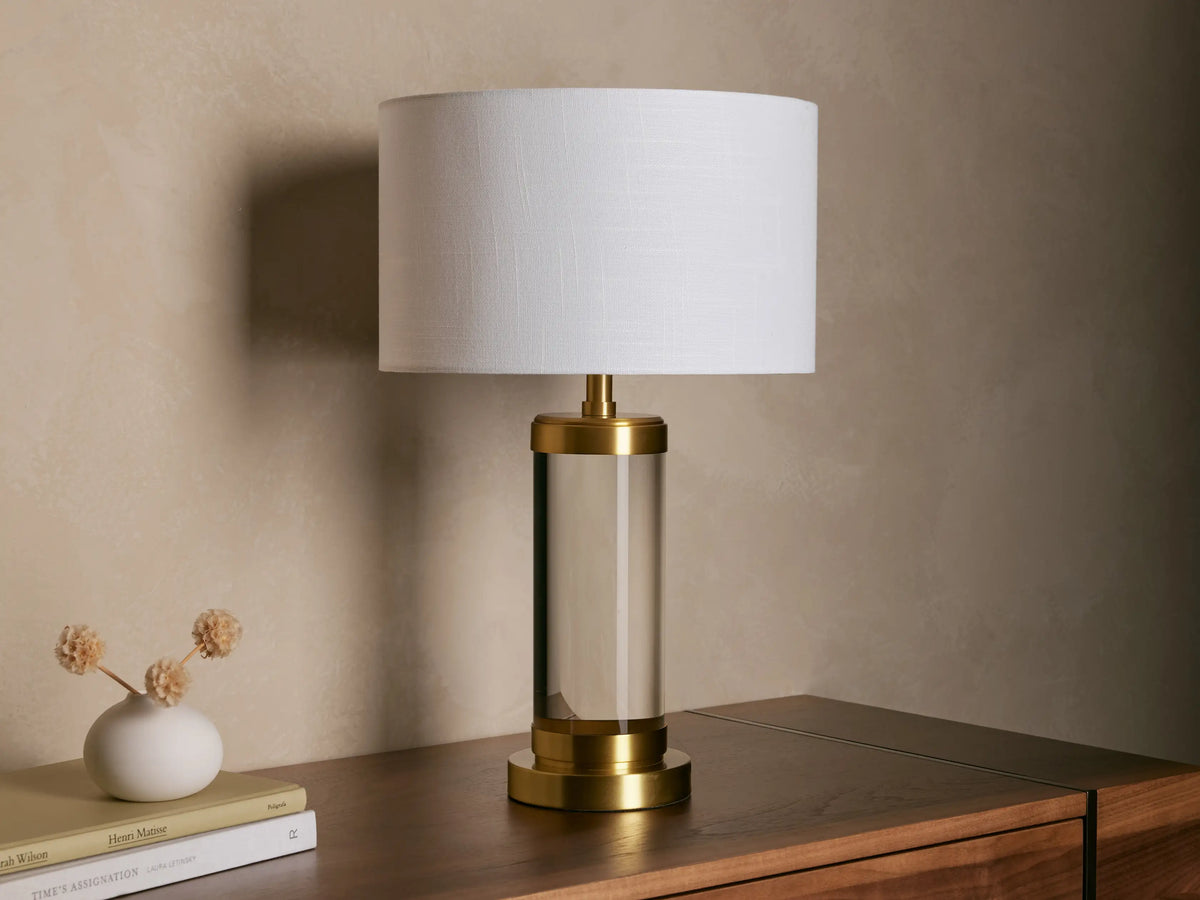 Linden Crystal Table Lamp with clear crystal column, brass accents, and off-white linen shade, glowing on a wooden table.