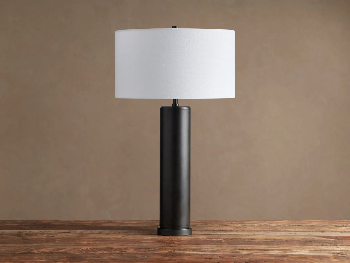 Linden Table Lamp with a dark cylindrical base, possibly pewter or bronze, and a white linen drum shade on a wooden surface.