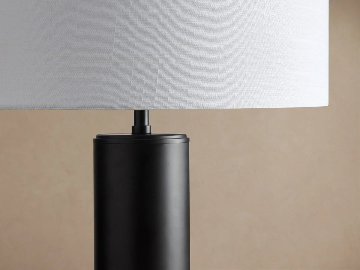 Linden Table Lamp with a dark cylindrical base and a white linen cylindrical shade, sitting on a rustic wooden table.