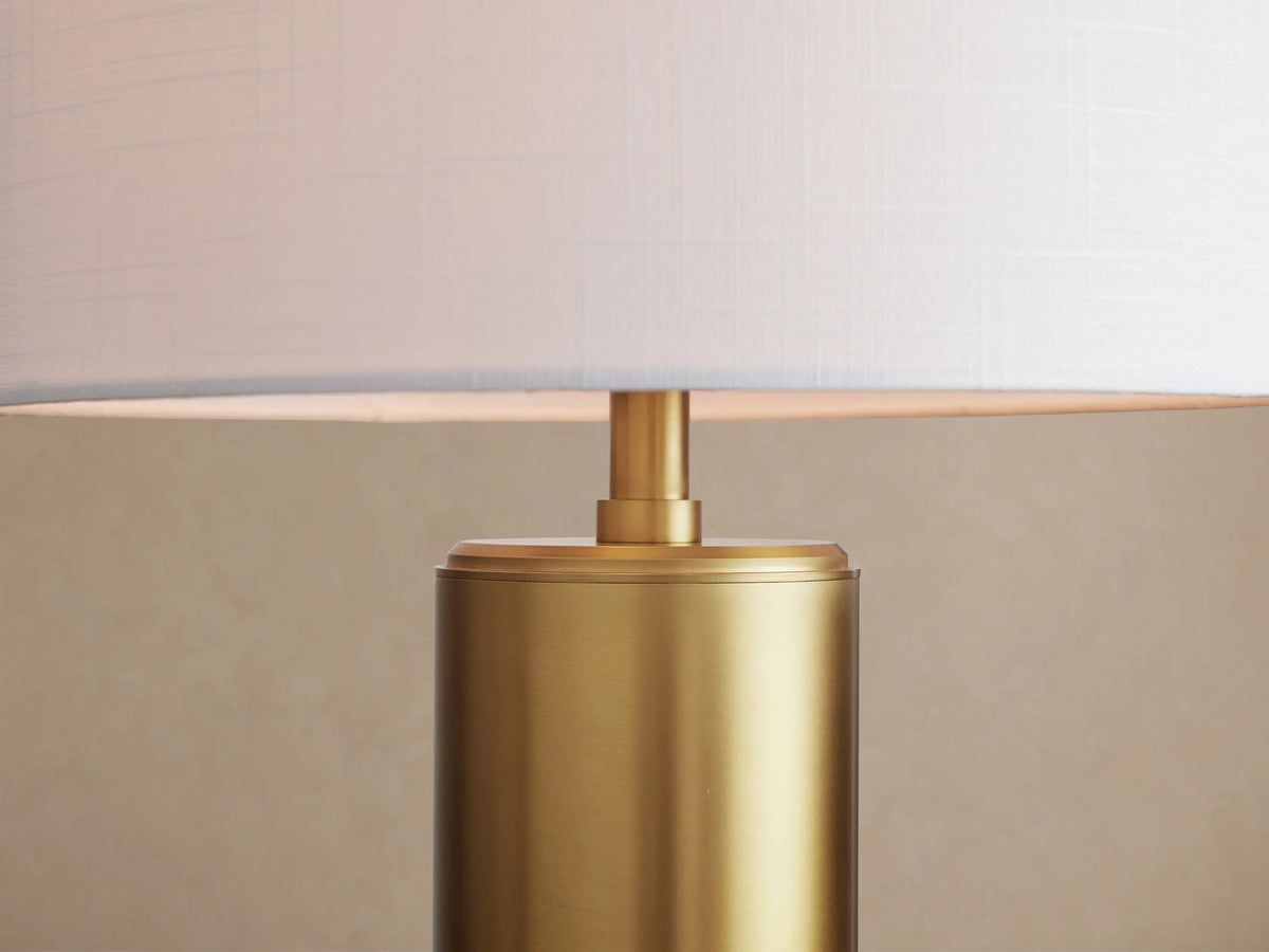 Linden Table Lamp with a cylindrical brass base and a white linen drum shade, sitting on a rustic wooden table.