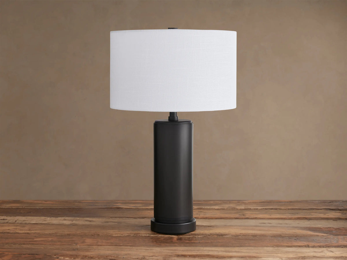 Linden Table Lamp: white linen shade on a dark cylindrical base with a bronze accent, centered on a rustic wooden table.