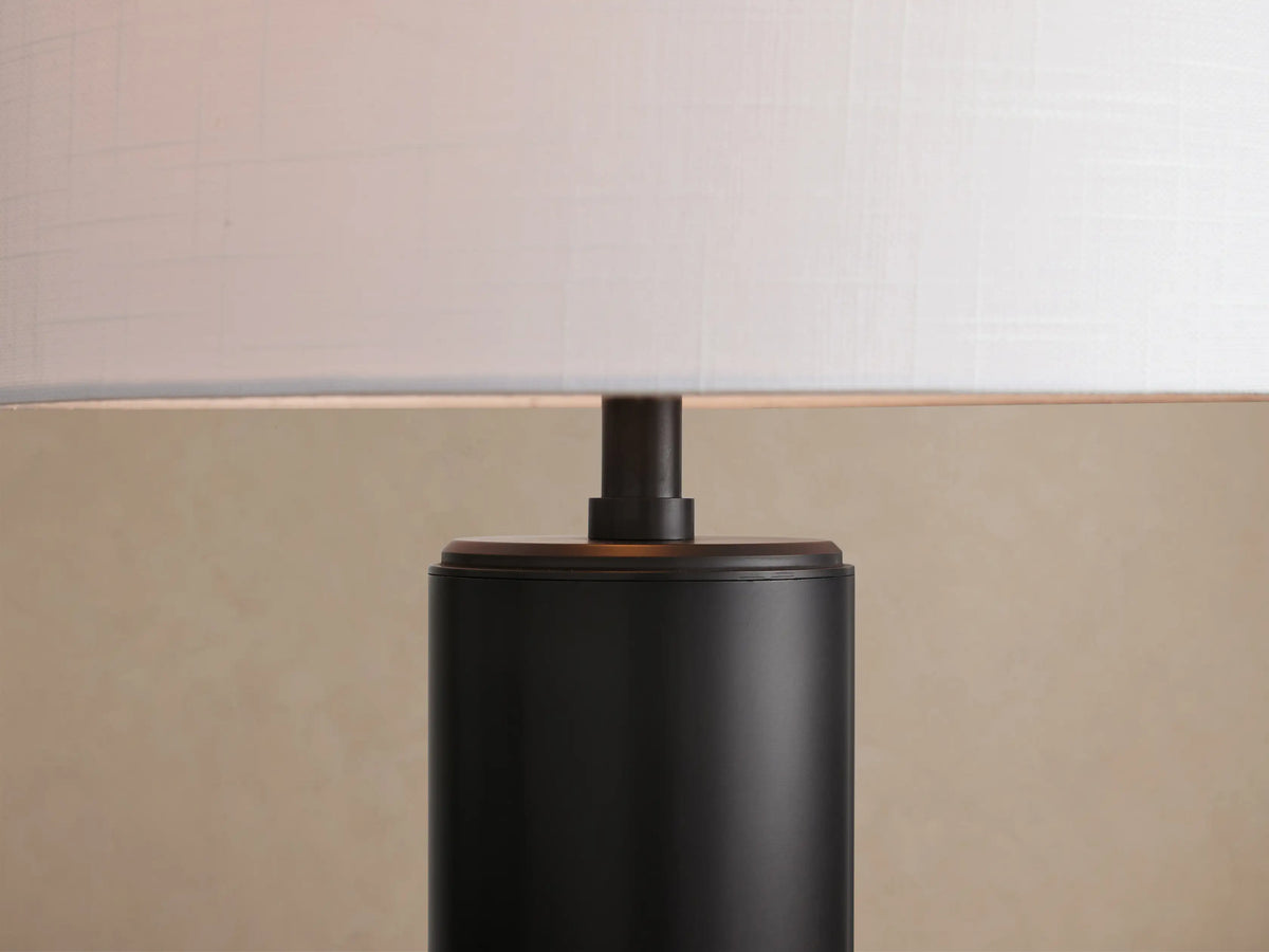 Linden Table Lamp with a black cylindrical base and a white linen drum shade, sitting on a rustic wooden table.