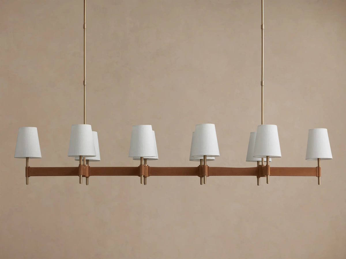 Hastings Linear Chandelier with brass structure, brown leather bar, six lit white linen shades, against a beige wall.
