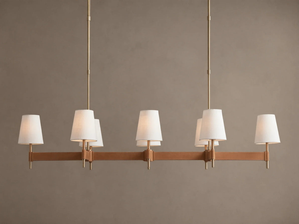 Hastings Linear Chandelier with 6 white linen shades, antiqued brass frame, and brown leather details on a plain wall.