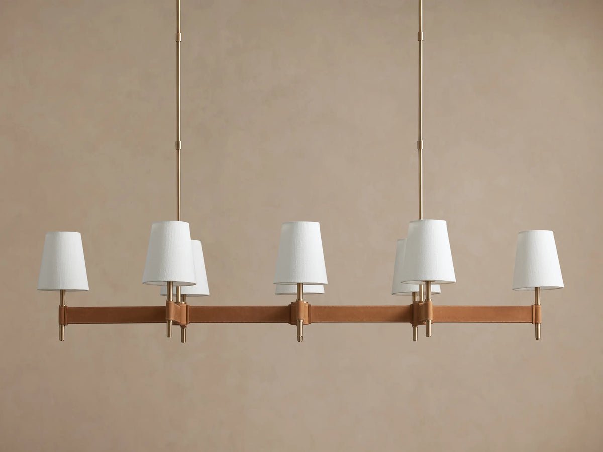 Hastings Linear Chandelier with 6 white linen shades, antiqued brass frame, and brown leather details on a plain wall.