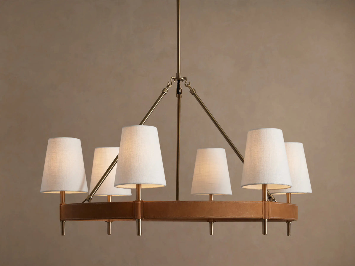 Round Hastings chandelier, 6 white linen shades emit warm light, with an antique brass frame & tan leather base.