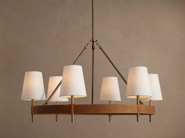 Round Hastings chandelier, 6 white linen shades emit warm light, with an antique brass frame & tan leather base.