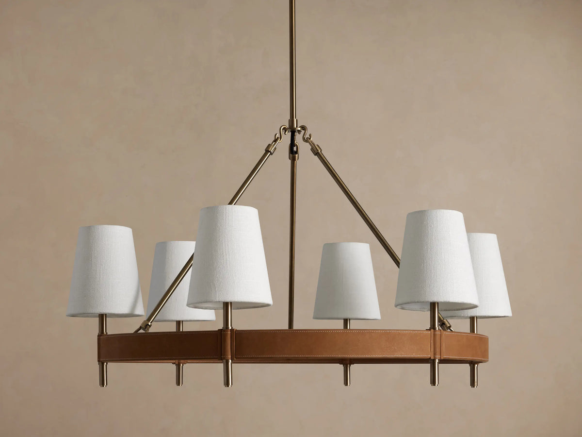 Round Hastings chandelier, 6 white linen shades emit warm light, with an antique brass frame & tan leather base.