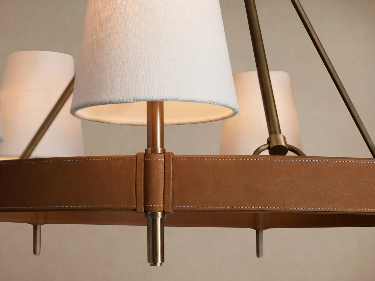 Hastings Round Chandelier with antiqued brass frame, brown leather band, and six white linen shades against a beige wall.