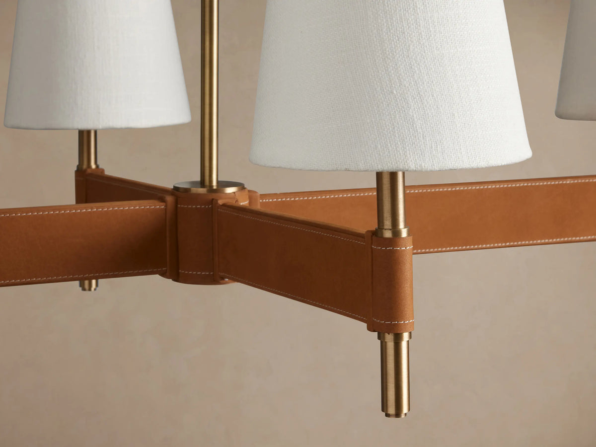Hastings Linear Chandelier with 6 white linen shades, antique brass, and brown leather bar, hanging against a beige wall.