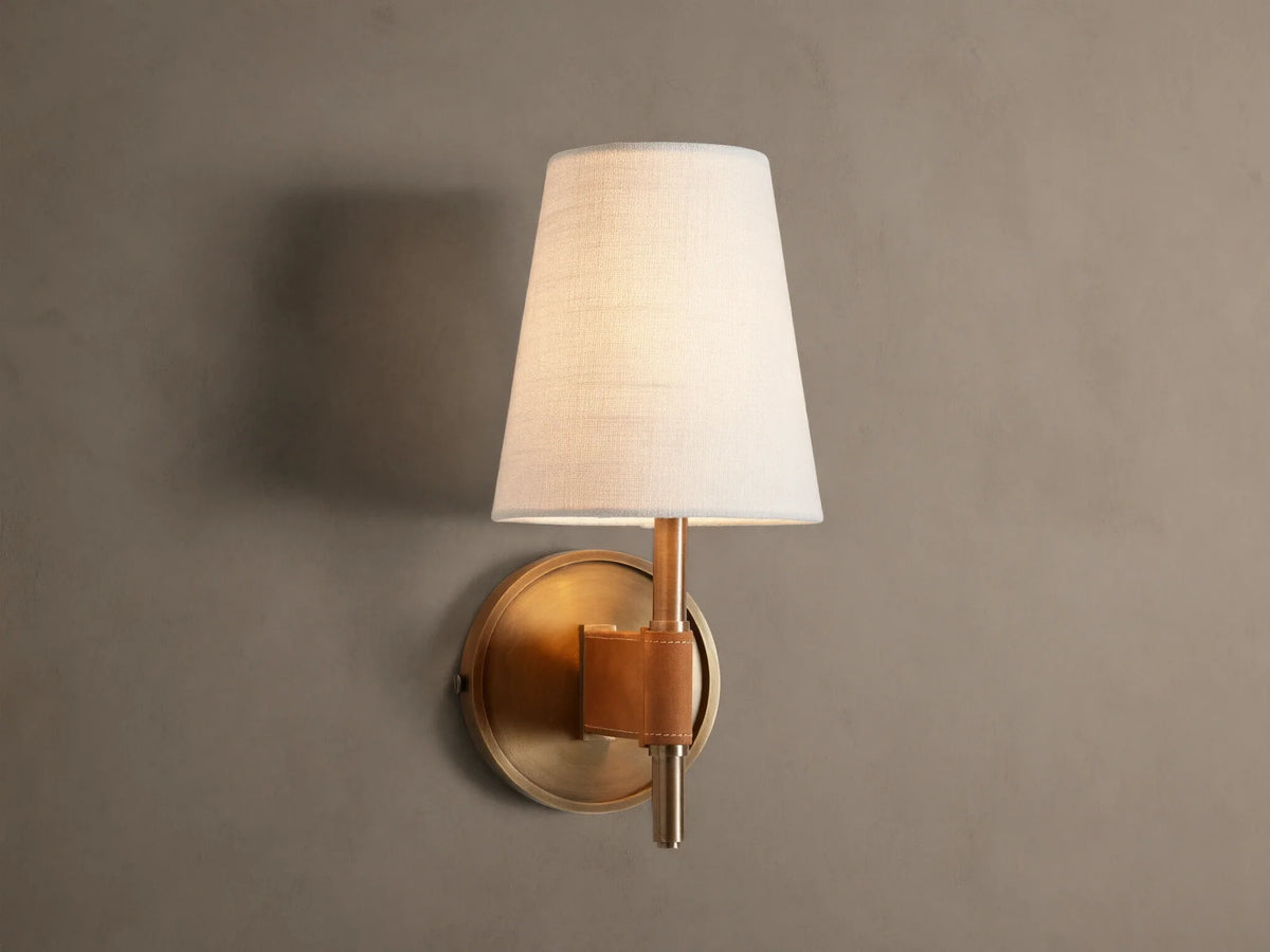 Hastings Sconce: Lit, antiqued brass wall sconce with a tapered white linen shade and brown leather accent, on a neutral wall.