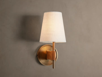 Hastings Sconce: Lit, antiqued brass wall sconce with a tapered white linen shade and brown leather accent, on a neutral wall.