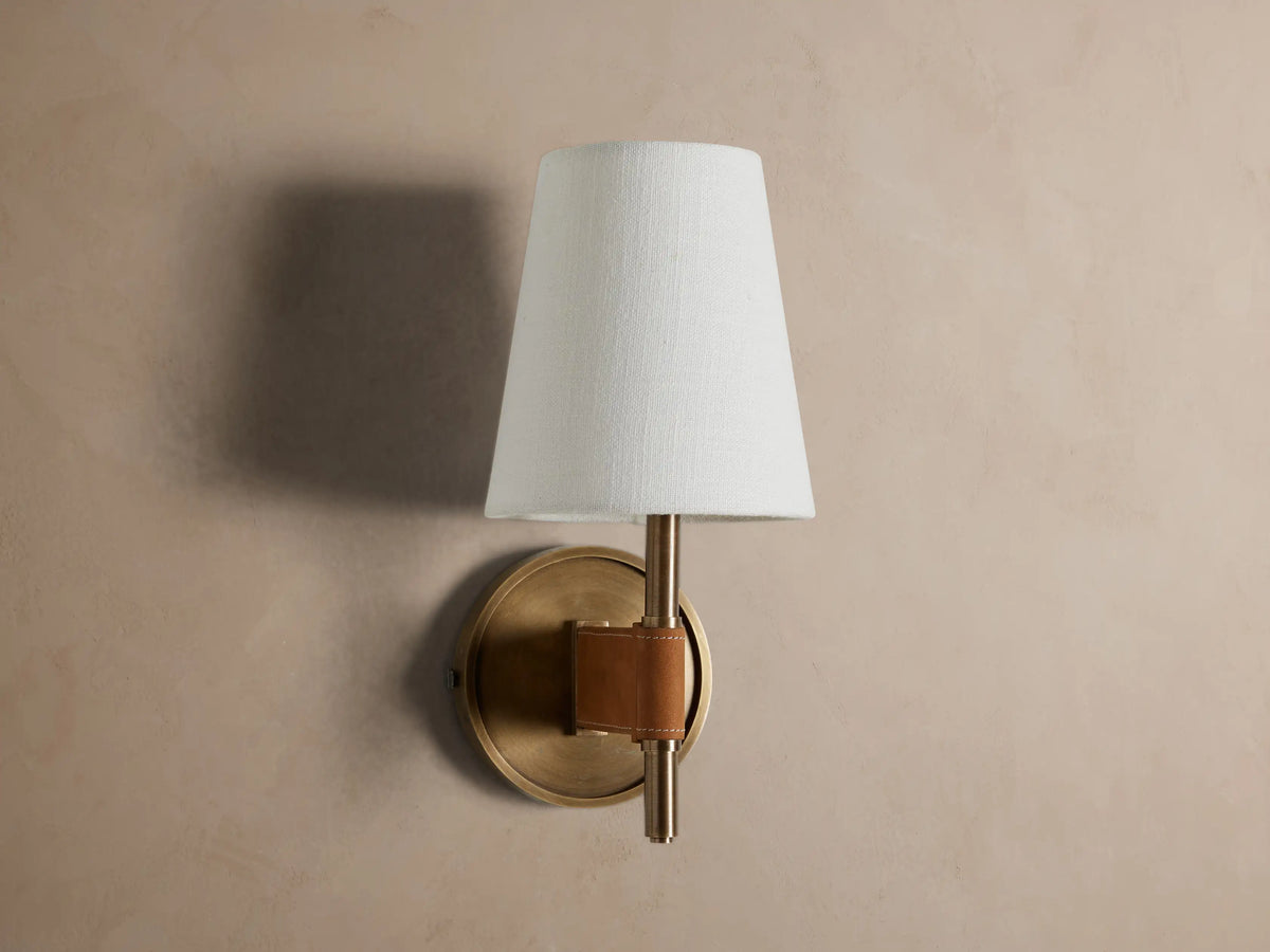 Hastings Sconce: Lit, antiqued brass wall sconce with a tapered white linen shade and brown leather accent, on a neutral wall.