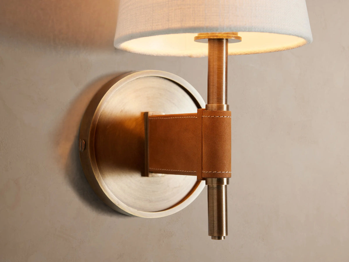 Hastings Sconce on a beige wall. It has an antiqued brass base, brown leather detail, and a white tapered linen shade.