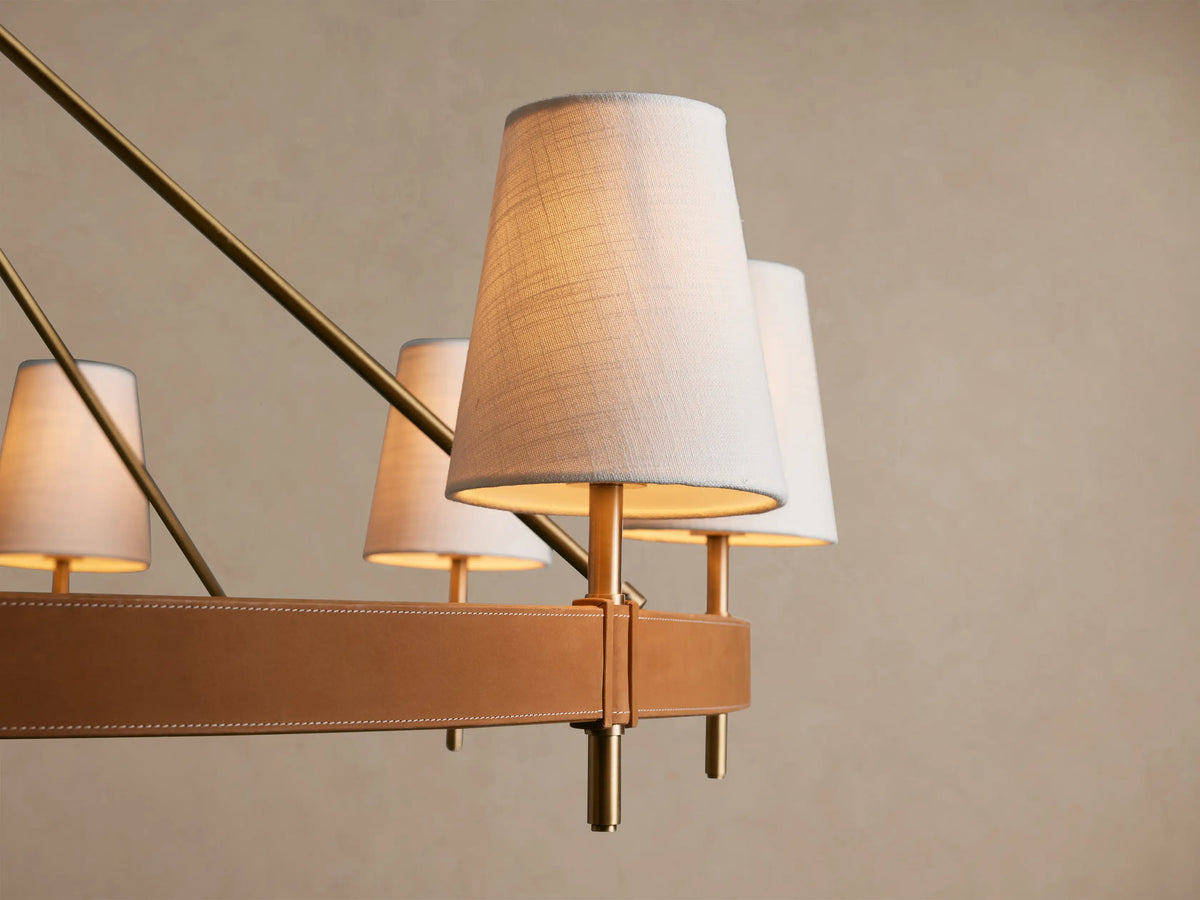 Round Hastings Chandelier with antiqued brass frame, brown leather details, and eight white linen shades, suspended.
